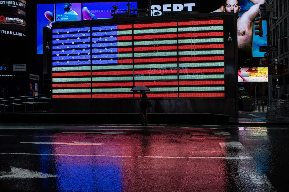 Lampoon, A lone man with an umbrella walks past the image of the United States flag on a huge screen in a nearly-empty Times Square. Hossein Fatemi Panos Pictures