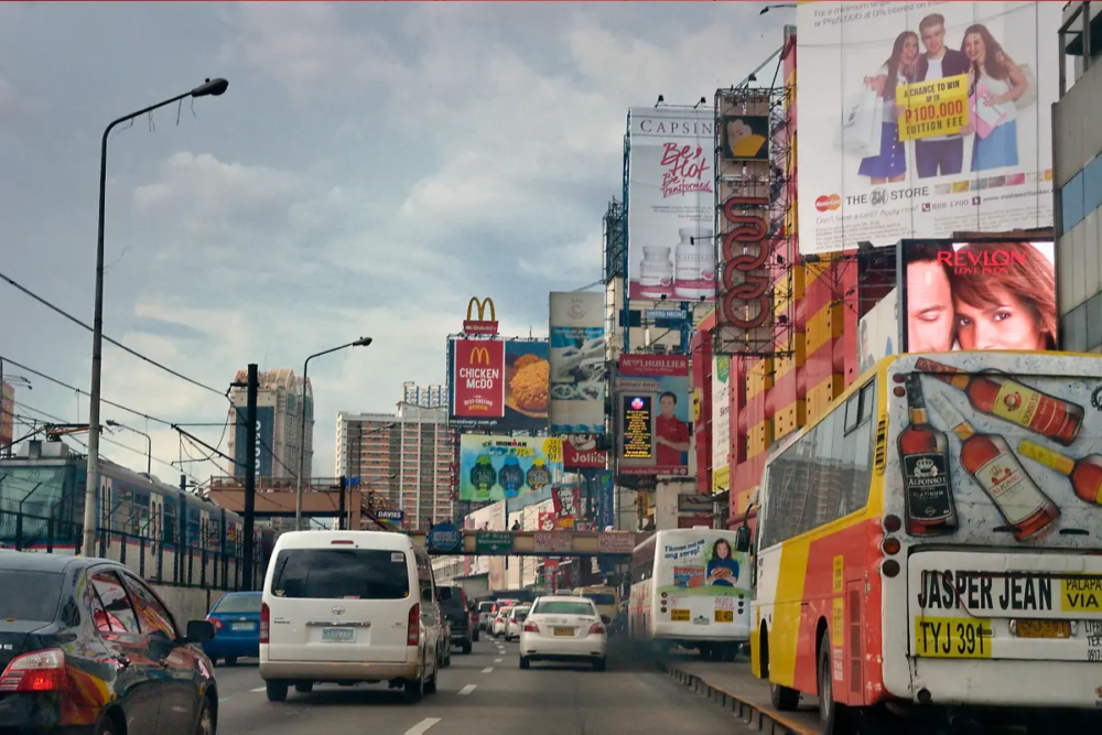 Advertising billboards hang from the side of buildings beside a busy road, Laurent Weyl Argos, Panos
