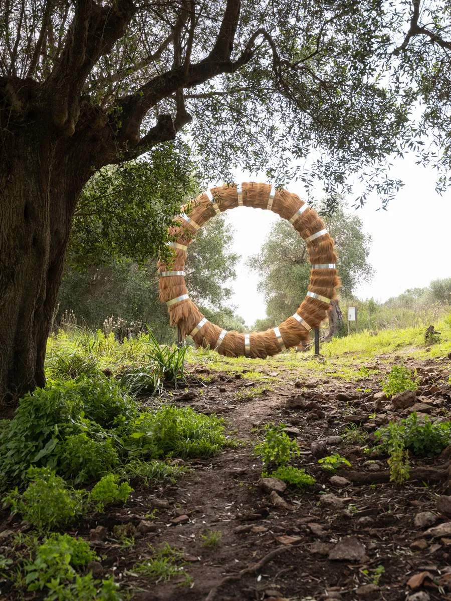 A large circular ring made of bundled reeds stands upright on two metal supports in a gallery. The ring frames a doorway behind it, creating a passage through its center. Metal bands wrap around the reeds at regular intervals, holding the structure together.