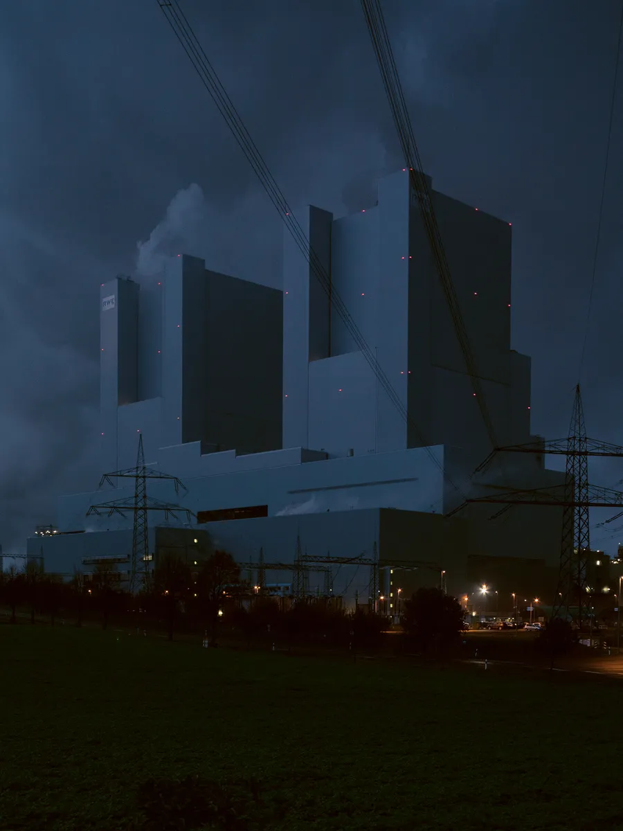 An industrial power plant with tall rectangular buildings rises against a dark, stormy sky, with transmission towers and power lines in the foreground.