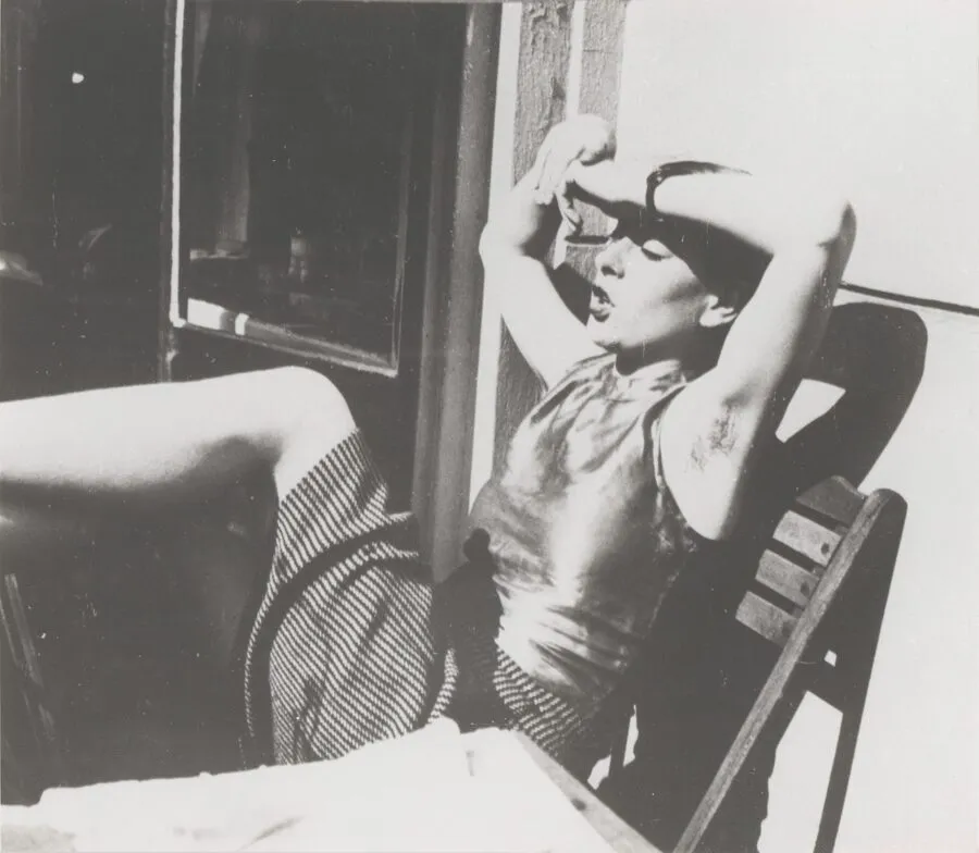 Black and white photograph of a woman leaning back in a wooden chair outdoors, shielding her eyes from bright sunlight with her arms raised.
