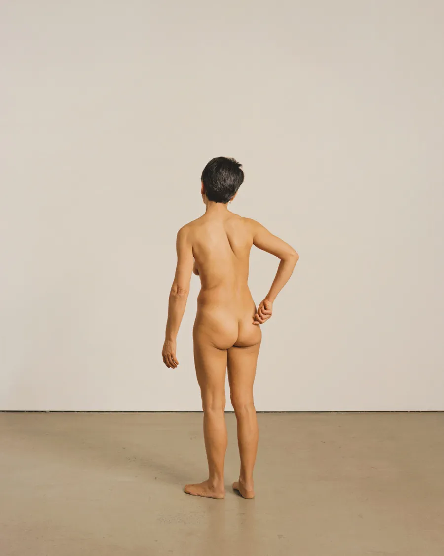 Rear view of a nude woman standing barefoot against a plain wall, one hand resting at her lower back.