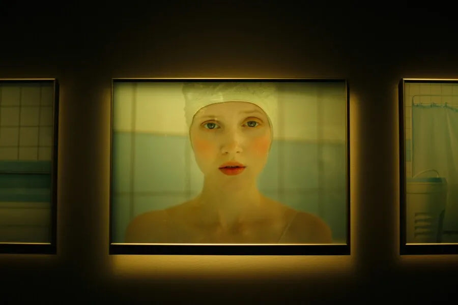Woman wearing a white swim cap, viewed through a softly lit glass panel in a green-tinted interior.