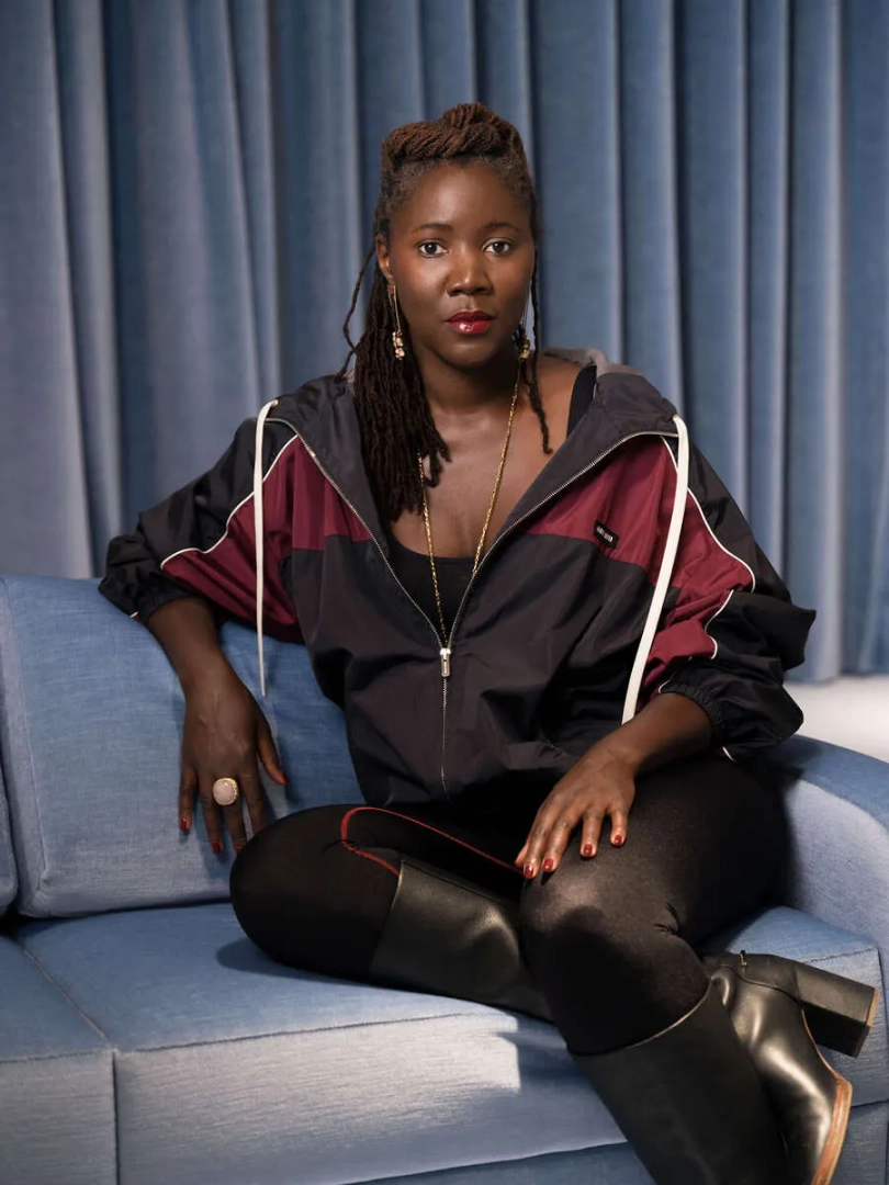 Portrait of filmmaker Alice Diop seated on a blue sofa, wearing a black and burgundy jacket, with blue curtains behind her.