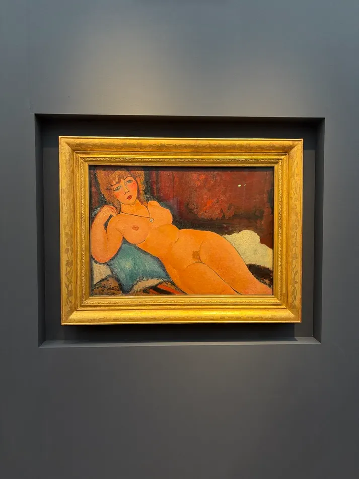 Reclining nude woman painted by Amedeo Modigliani, with warm orange skin tones, dark hair and a blue cushion, displayed in an ornate gold frame.