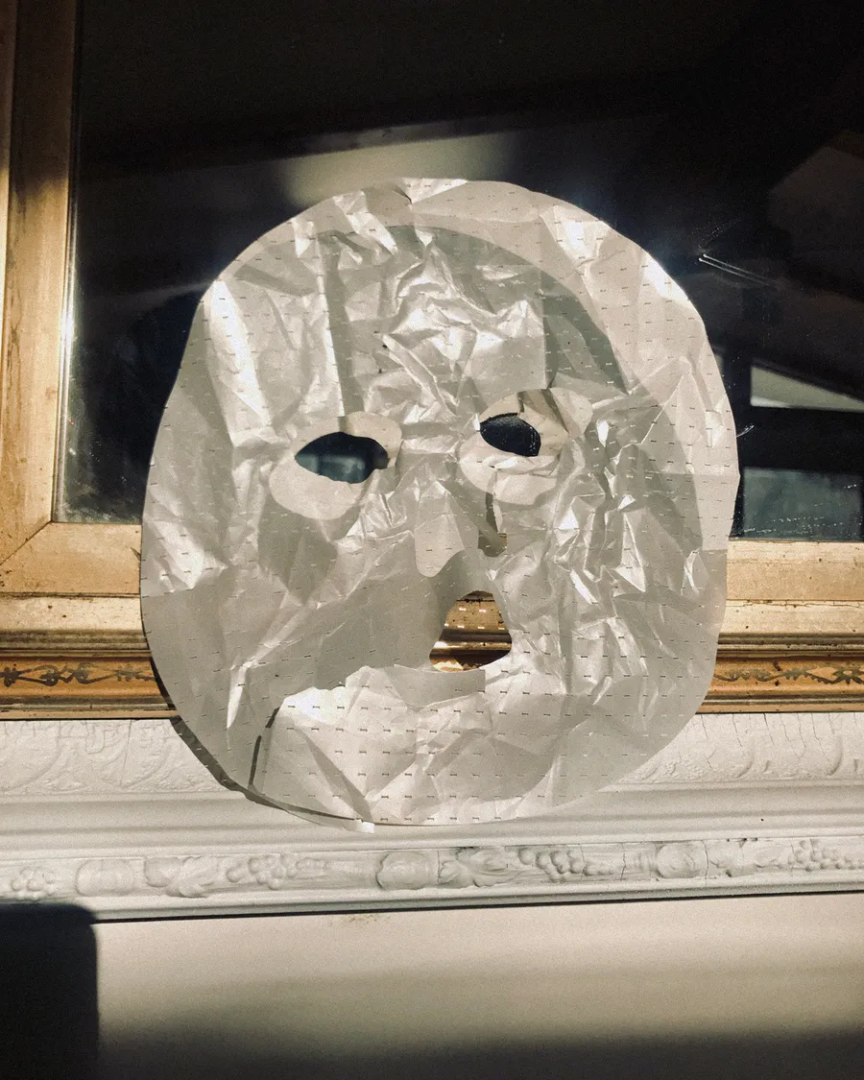 Wrinkled white sheet mask with cutouts for the eyes, nose, and mouth displayed against a dark window above an ornate white mantelpiece.