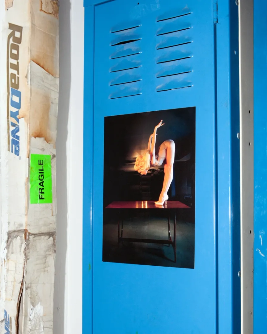 Photograph of a blonde performer bending backward on a table, attached to a blue metal locker beside a green fragile sticker.