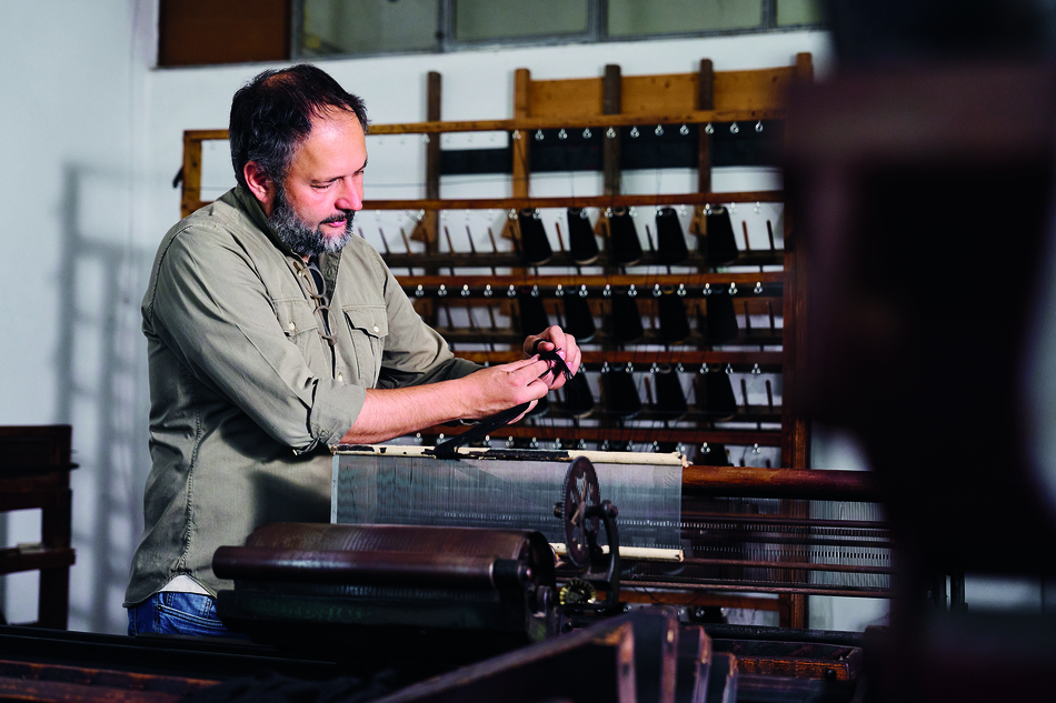 Carlo Colombo working at a traditional wooden weaving loom, adjusting threads by hand inside a textile workshop.