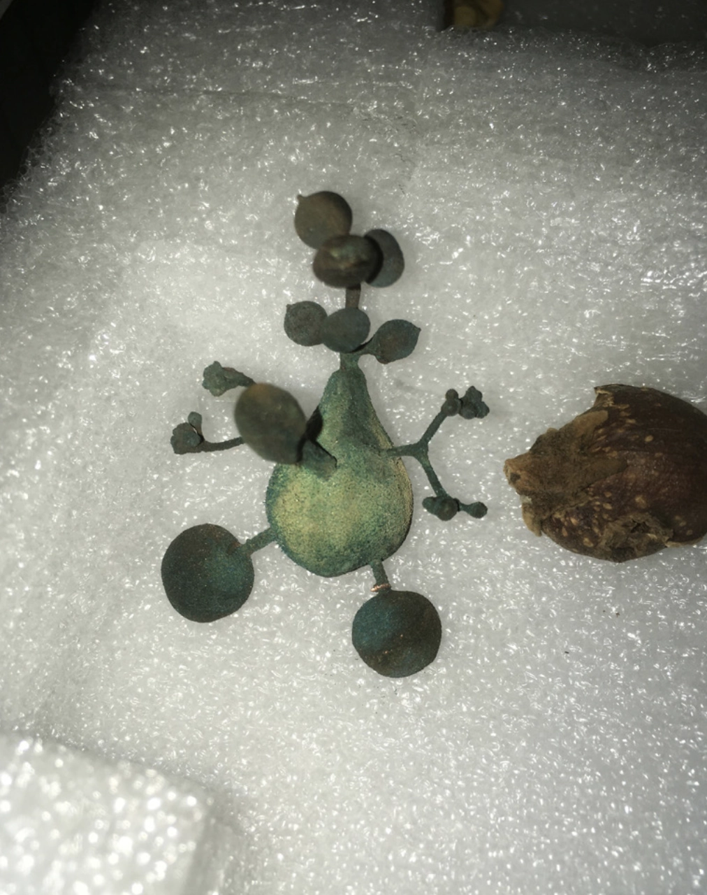 Green pewter Daphne sculpture with branching limbs and rounded forms resting on white foam beside a dried root.