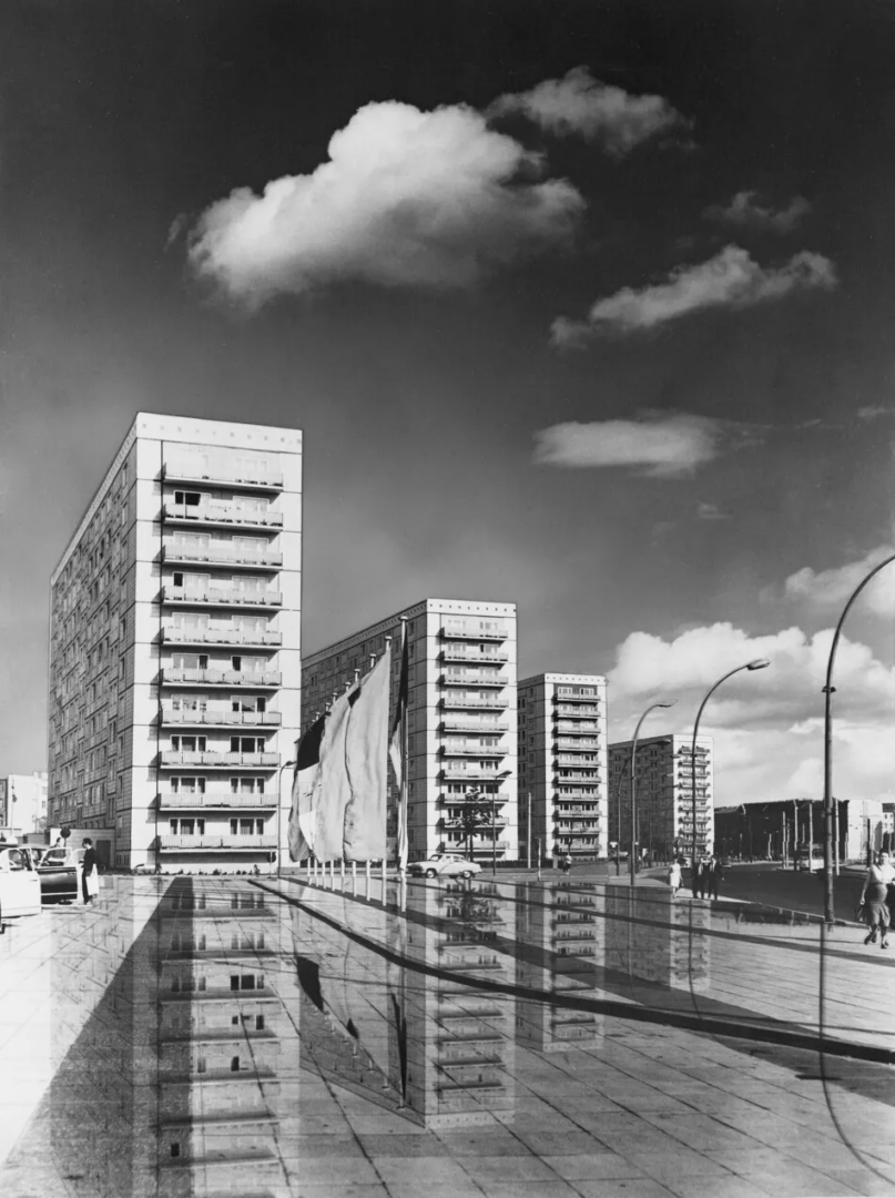 Black-and-white architectural collage of prefabricated residential towers on Alexanderstraße in East Berlin, with balconies, streetlights and reflections across a paved foreground.