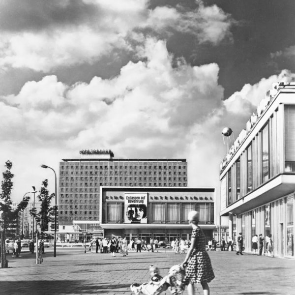 Black-and-white photograph of Hotel Berolina and Kino International in East Berlin in 1966, with pedestrians, a woman pushing a pram and modernist buildings beneath a cloudy sky.