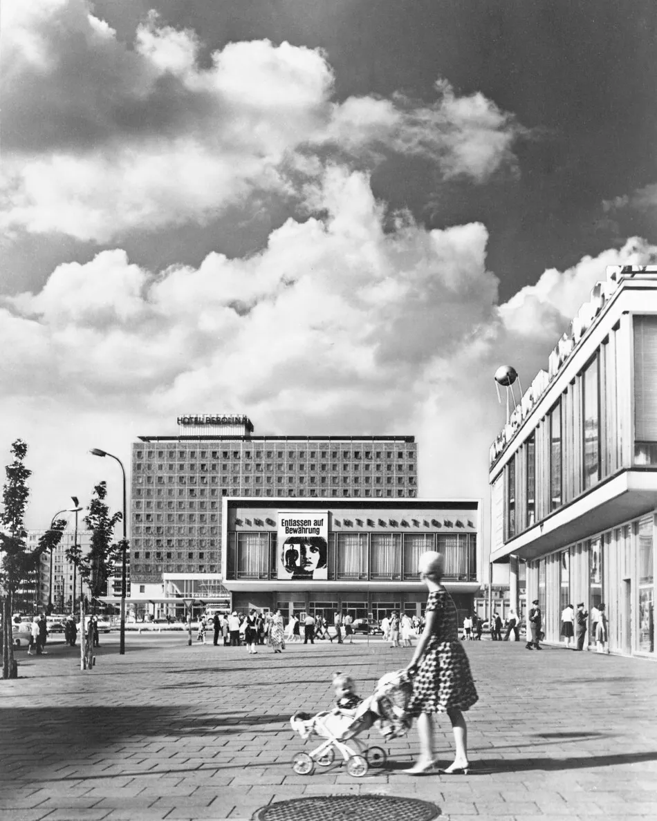 Black-and-white photograph of Hotel Berolina and Kino International in East Berlin in 1966, with pedestrians, a woman pushing a pram and modernist buildings beneath a cloudy sky.