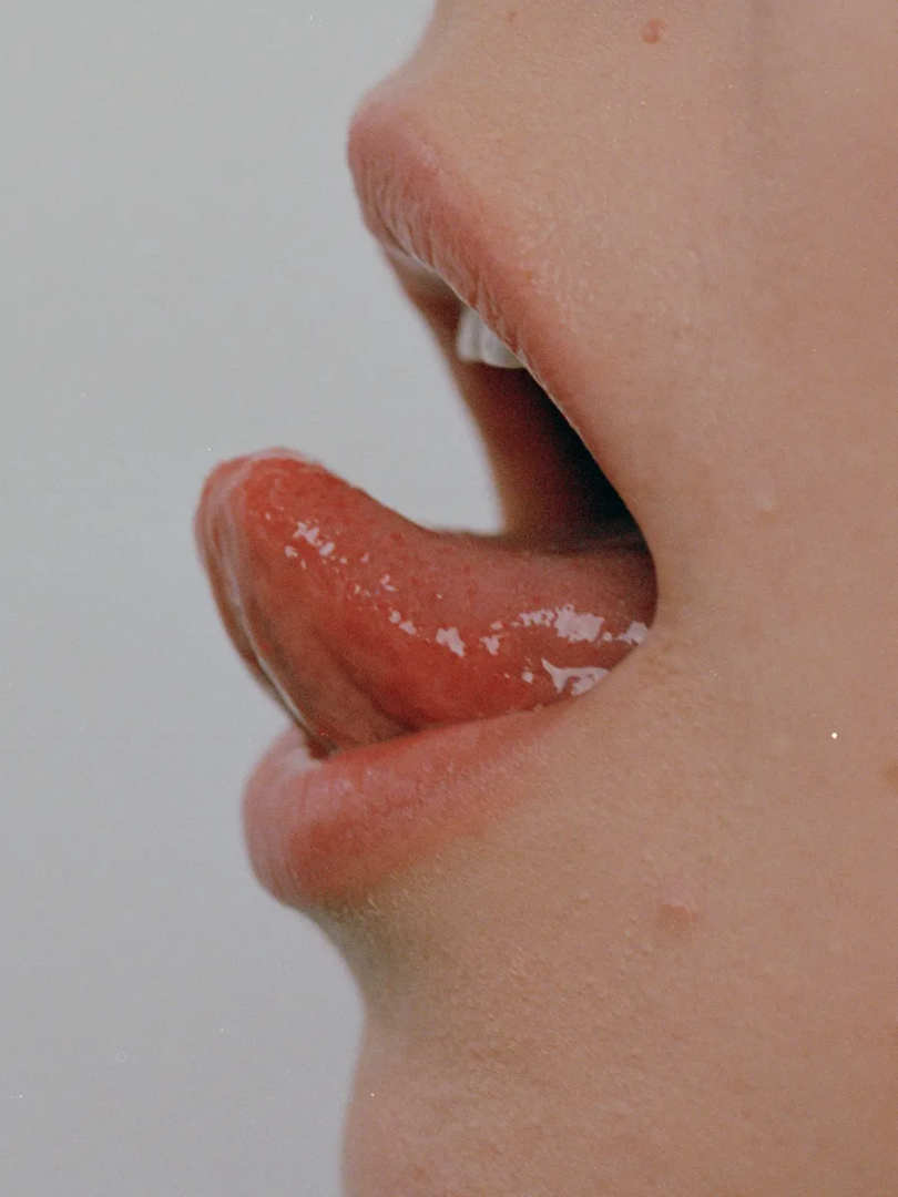 Side-profile macro photograph of an open mouth with a wet tongue extending outward against a pale background.