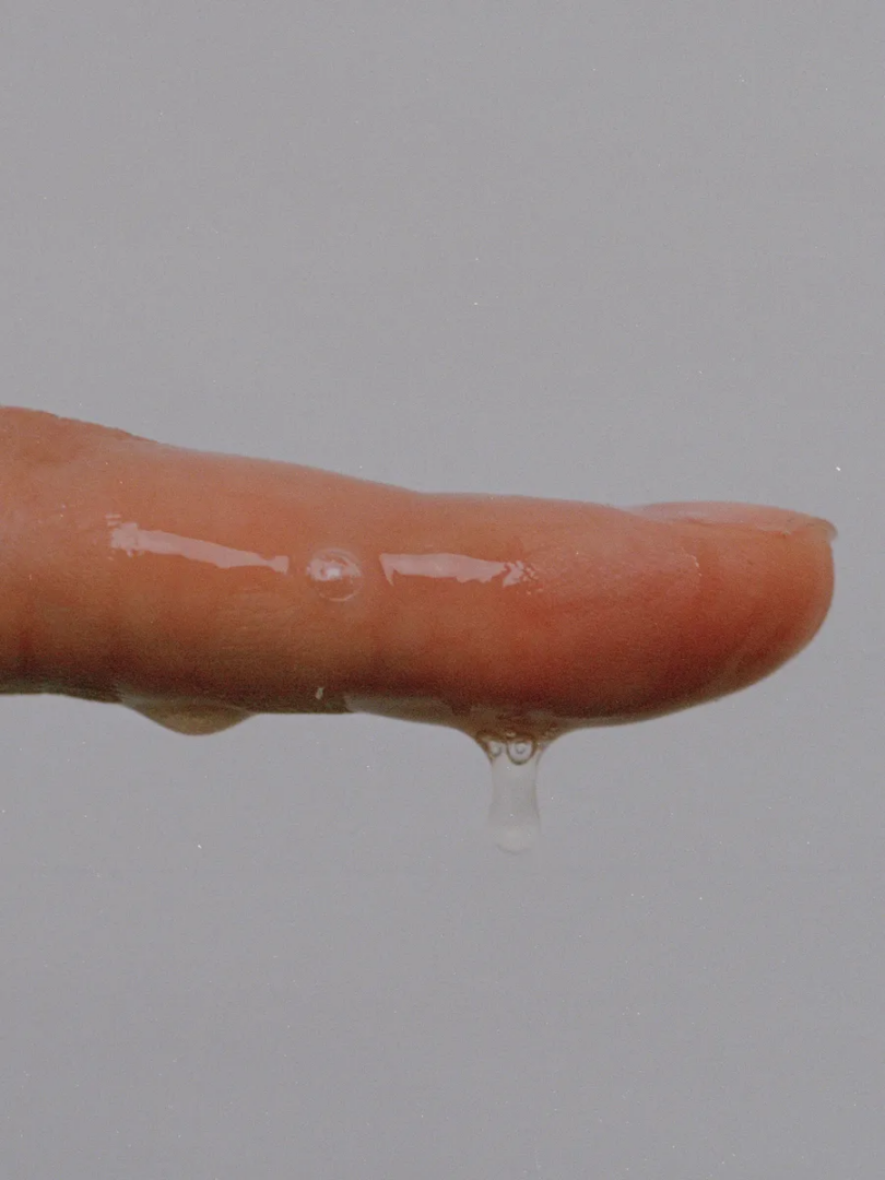Macro photograph of a saliva-coated finger with transparent droplets suspended beneath it against a grey background.