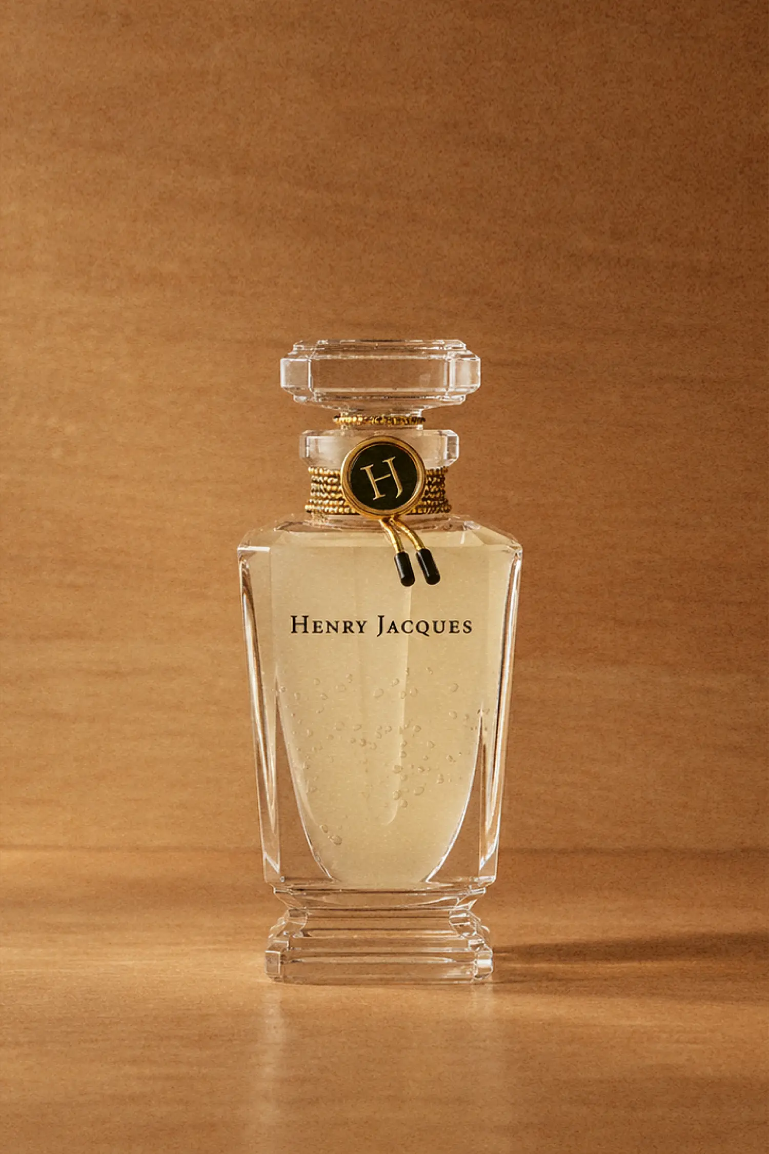 Henry Jacques Rose Snow de HJ perfume in a clear crystal flacon containing pale yellow concentrate with visible particles, set against a warm brown background.