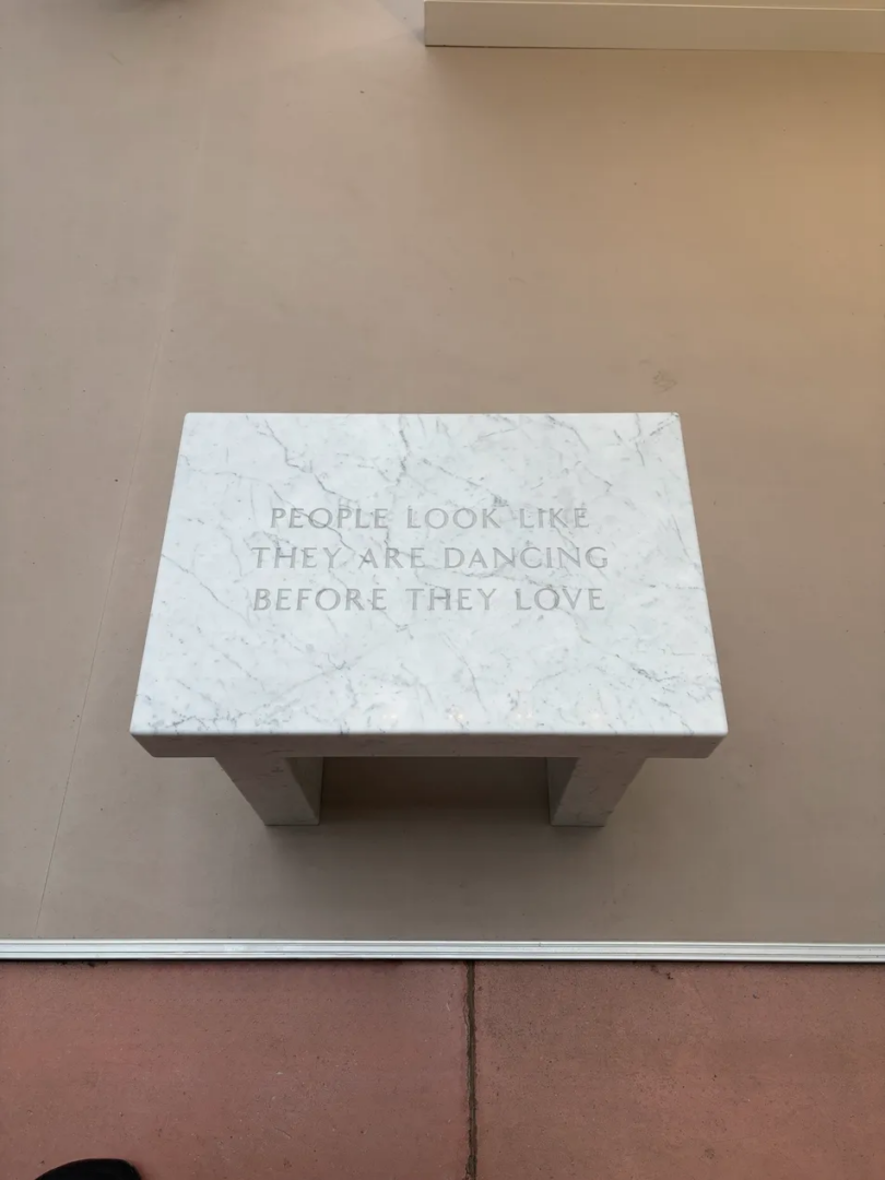 White marble bench by Jenny Holzer engraved with the words “People look like they are dancing before they love,” displayed on a pale gallery floor.
