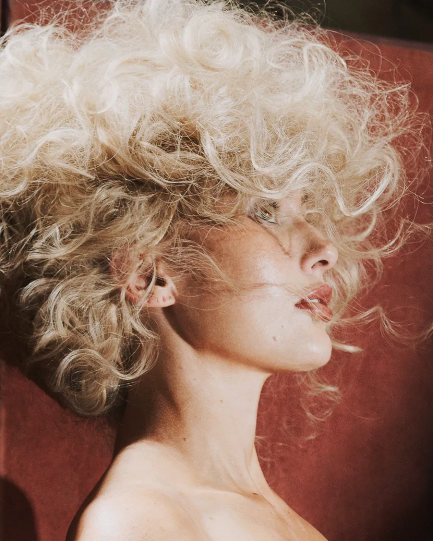 Artistic side-profile portrait of Maja Malou Lyse with voluminous platinum-blonde curls, soft makeup and a warm red background, photographed by Lalo + Eva