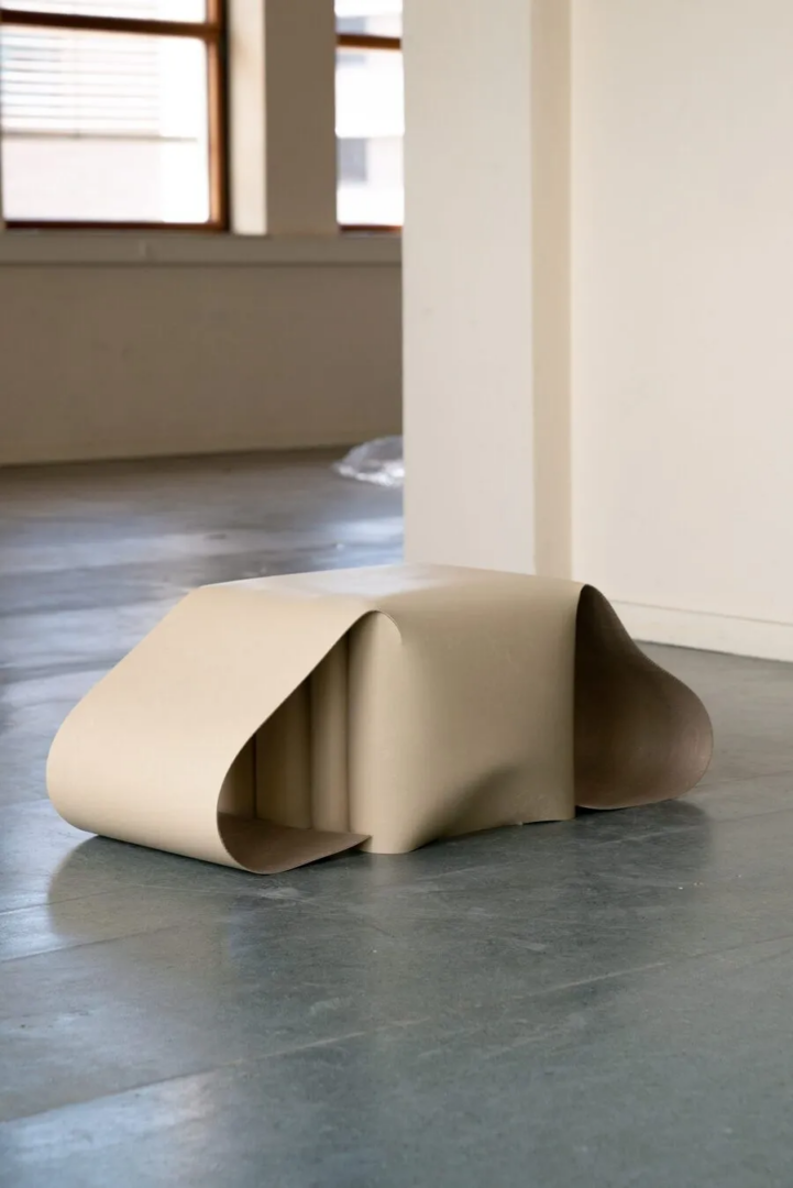 Sculptural linoleum bench with a folded beige surface and curved supports, displayed on a concrete floor.