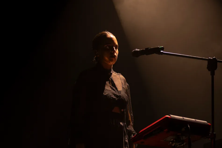 Close-up of Marina Herlop standing beside a microphone and electronic percussion instrument, her face illuminated by amber stage lighting against a dark background.