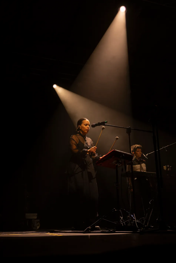 Marina Herlop plays an electronic percussion instrument with two mallets beneath converging spotlights, accompanied by another musician on a dark stage at Unsound Festival.