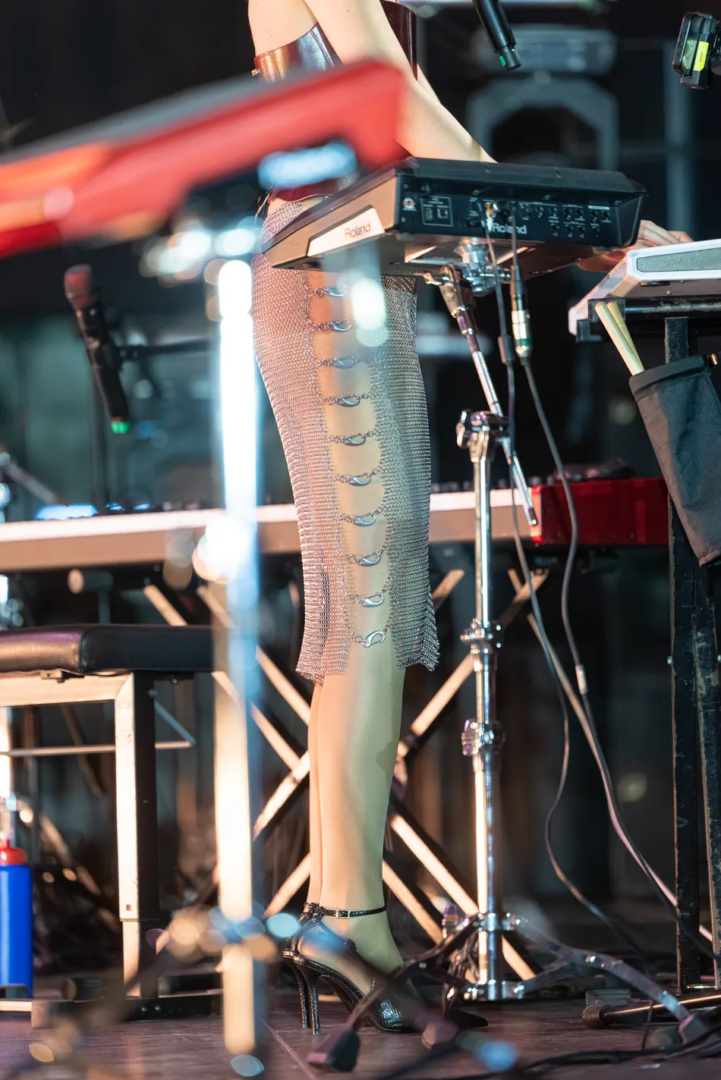 Detail of Marina Herlop's stage outfit, featuring a metallic mesh skirt with silver fastenings and black high heels, photographed beside electronic keyboards, microphones and cables.