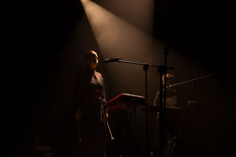 Close-up of Marina Herlop standing beside a microphone and electronic percussion instrument, her face illuminated by amber stage lighting against a dark background.