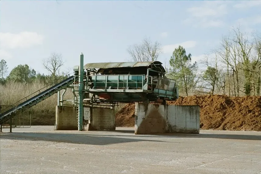 Industrial conveyor belt and rotating screening machinery at an outdoor waste processing facility, with piles of organic material.