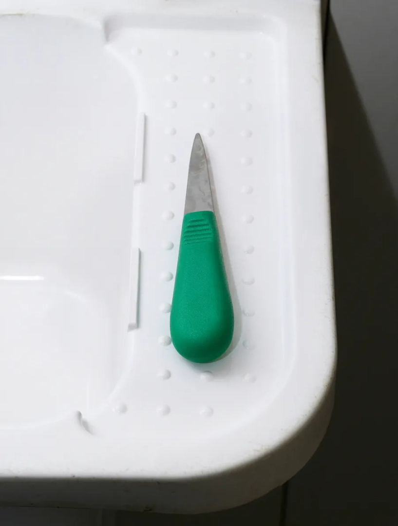 Close-up of an oyster shucking knife with a green handle and stainless steel blade, resting on a white textured plastic surface