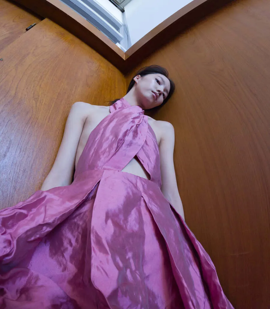 Miu Miu SS27 pink satin halter dress with a cutout bodice, photographed from a low angle against wood-paneled walls.