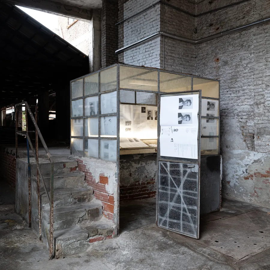 Paint It Black Torino exhibition space, a glass and metal installation displaying archival photographs and printed material against exposed brick walls.