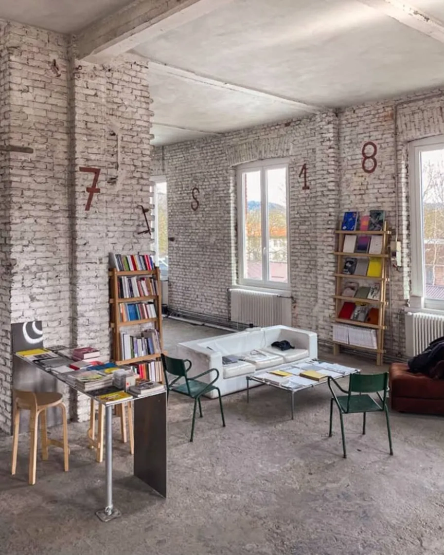 Paint It Black Torino reading area, with exposed brick walls, numbered columns, bookshelves, a white sofa and metal chairs arranged around low tables.