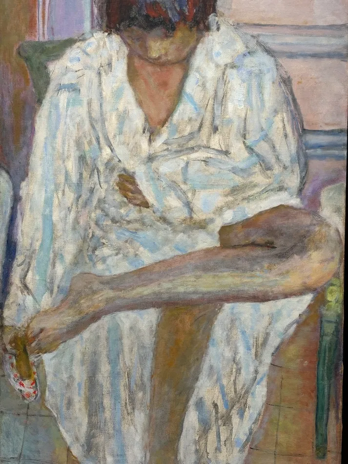 Intimate painting by Pierre Bonnard depicting a seated woman in a loose white robe, bending forward with one bare leg raised across her lap, rendered in muted blue, pink and cream tones.