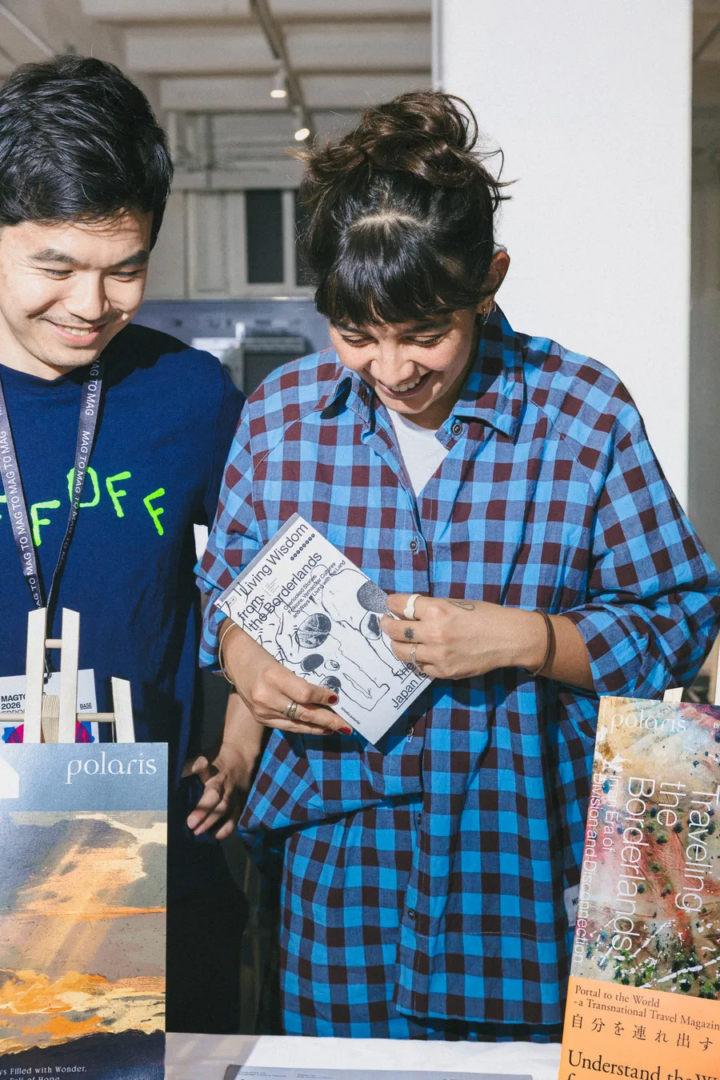 Visitors browsing magazines and independent publications across exhibitor tables inside the Mag to Mag 2026 fair, beneath an industrial ceiling and large windows.