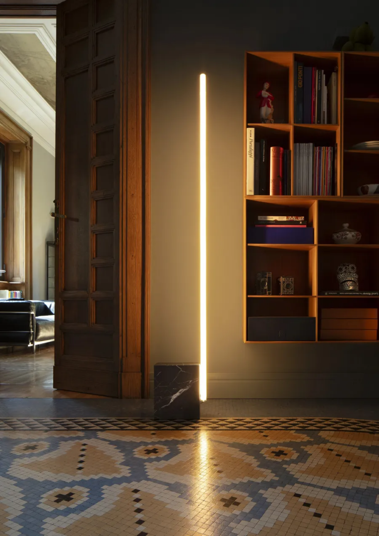 Flos Relay floor lamp designed by Michael Anastassiades, featuring a vertical LED light and dark marble base, displayed beside a wooden bookshelf in an elegant interior with patterned mosaic flooring