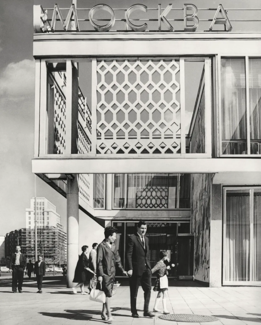 Black-and-white street photograph of Restaurant Moskau on Karl-Marx-Allee in East Berlin, showing its geometric facade, rooftop lettering and pedestrians outside the entrance.