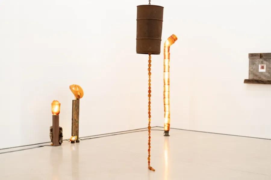 Sculptural installation featuring suspended cylindrical forms, hanging amber beads, and illuminated orange vertical structures inside a white gallery.