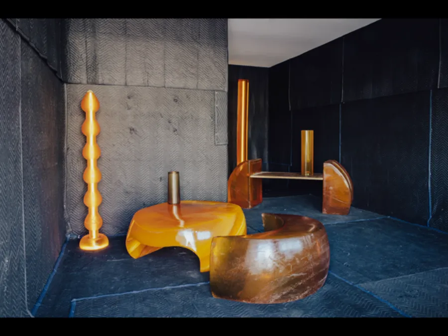 Installation of amber and brown sculptural furniture, including curved seating, a low table, and vertical forms, arranged inside a dark, padded room.