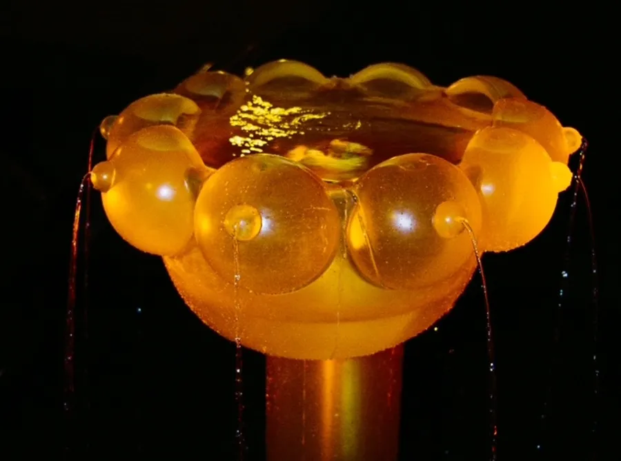 Glowing amber sculpture with rounded protrusions and thin streams of liquid dripping from its edges against a black background