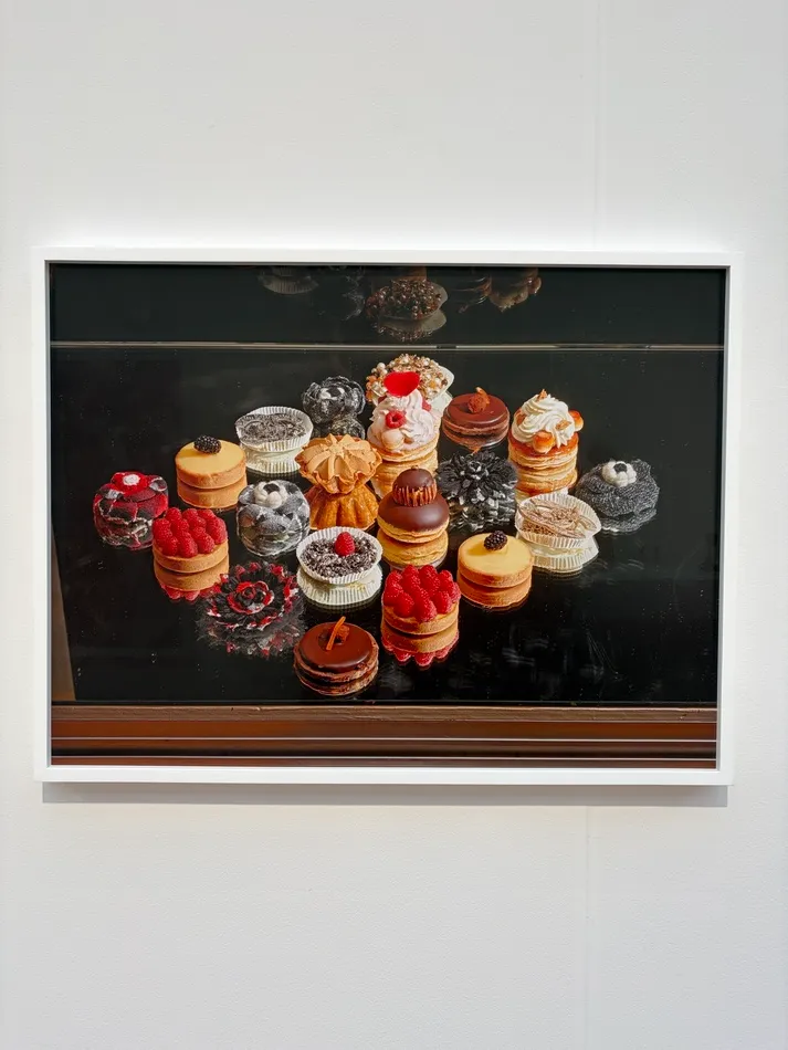 Color photograph by Roe Ethridge showing an assortment of elaborate miniature cakes and pastries decorated with raspberries, chocolate, cream and fruit on a reflective black surface.