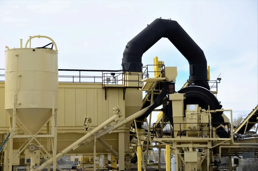 Industrial processing plant with large cylindrical storage silo, mechanical conveyors and black ventilation ducts.