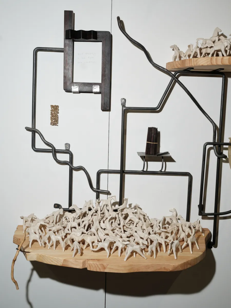 Installation of bent black metal rods and wooden shelves supporting groups of miniature white horses, with a dark wooden frame mounted above.