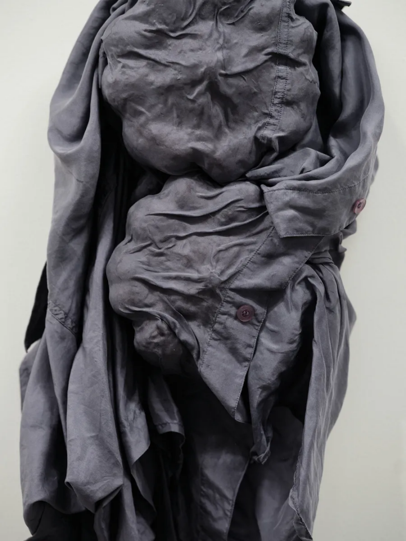 Close-up of a grey fabric sculpture made from gathered garments, showing wrinkles, seams, buttons and overlapping layers.