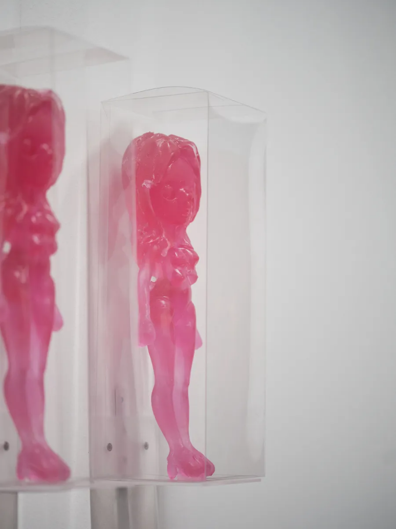 Two translucent pink figurines with elongated bodies and sculpted hair displayed inside transparent rectangular cases.