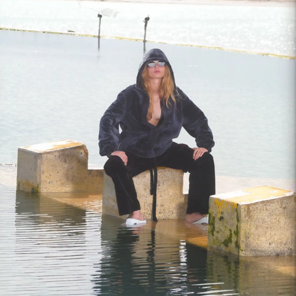 Self Service Issue 65 fashion editorial featuring Ida seated on concrete blocks by the water, wearing a dark hooded jacket and trousers by Phoebe Philo with sunglasses and archive shoes.