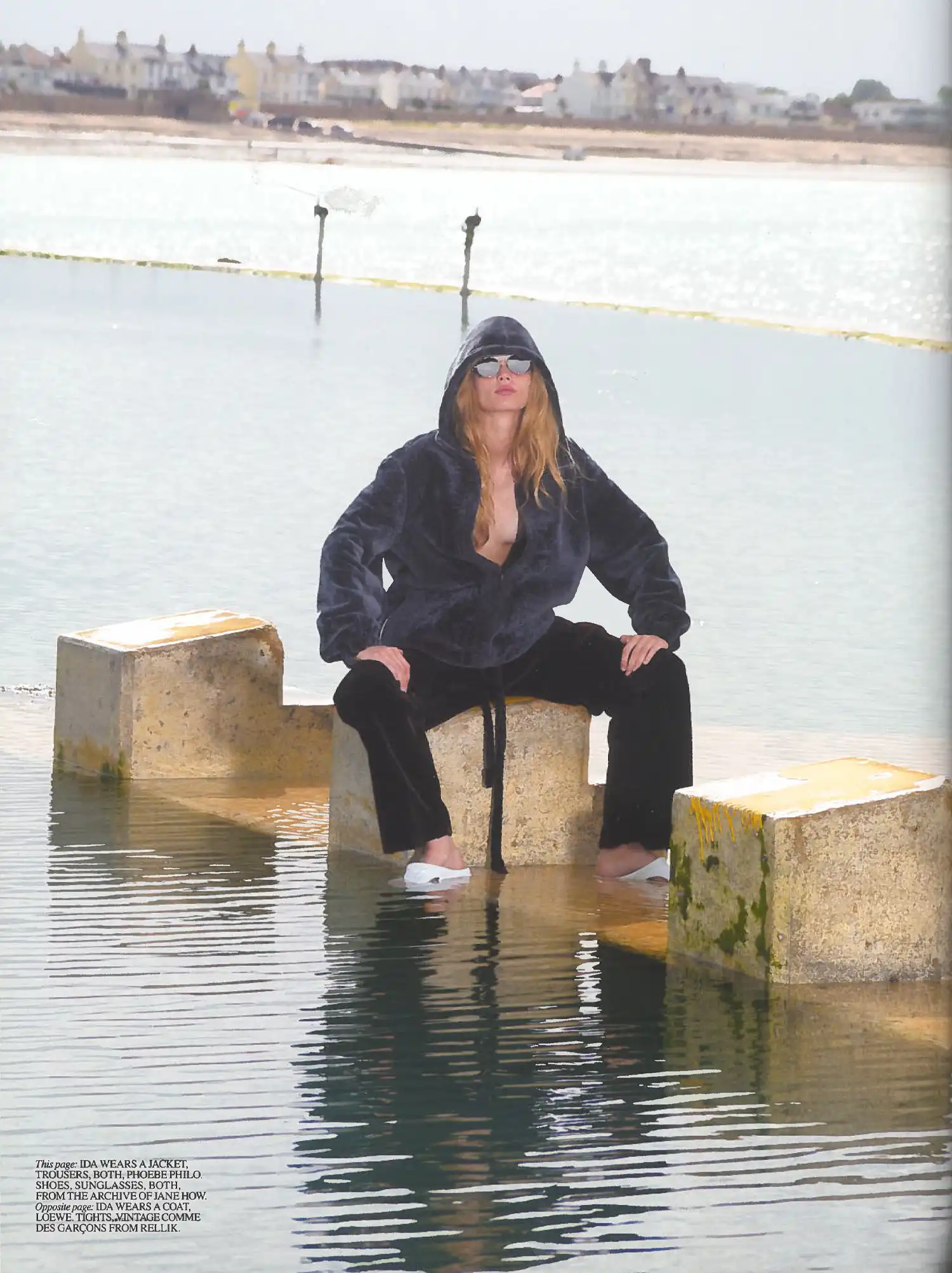 Self Service Issue 65 fashion editorial featuring Ida seated on concrete blocks by the water, wearing a dark hooded jacket and trousers by Phoebe Philo with sunglasses and archive shoes.