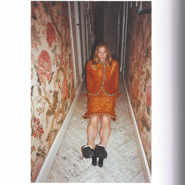 Self Service Issue 65 Chanel fashion editorial featuring a model in an orange textured skirt suit seated on the floor of a narrow hallway lined with floral tapestry.