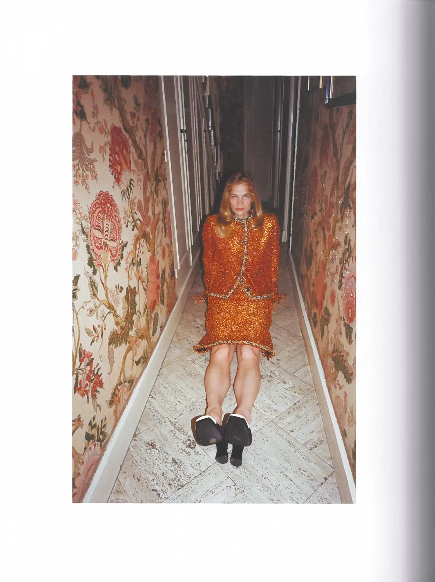Self Service Issue 65 Chanel fashion editorial featuring a model in an orange textured skirt suit seated on the floor of a narrow hallway lined with floral tapestry.