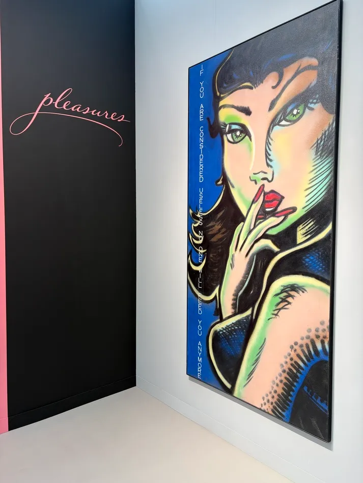 Gallery installation featuring a large Pop Art-style portrait of a woman with green eyes and red lips, her fingers raised to her mouth, alongside pink cursive lettering reading “pleasures” on a black wall.