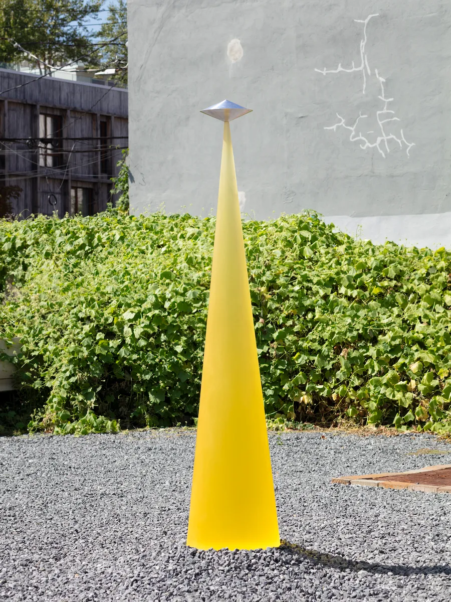 Tall yellow conical sculpture topped with a silver geometric form, standing on gravel in front of green vegetation.