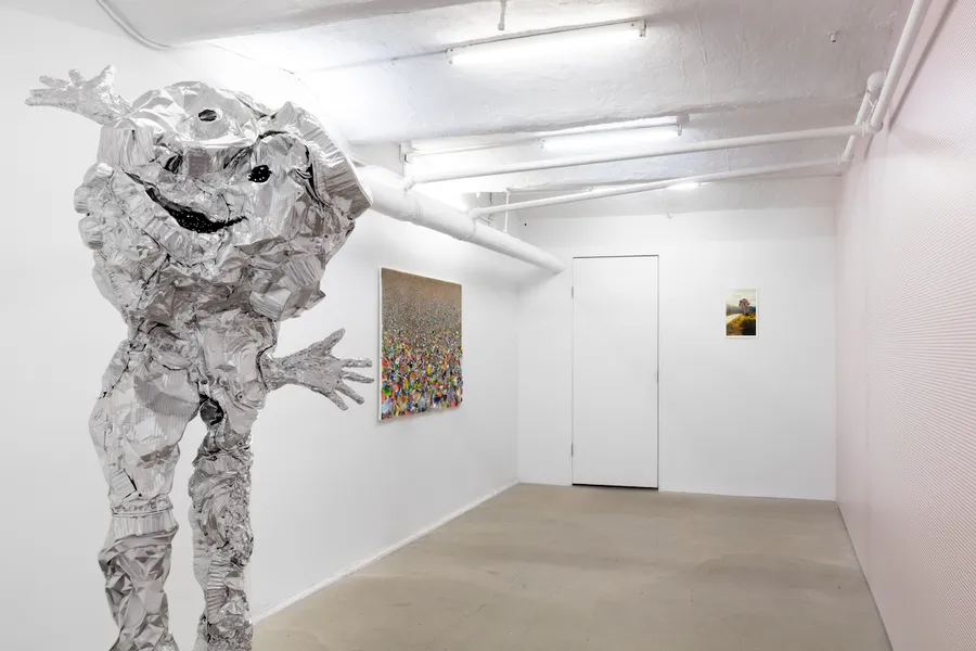 Interior gallery view featuring a silver human-like sculpture with outstretched arms and two artworks mounted on white walls.